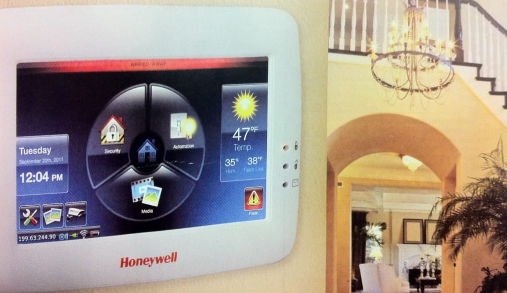 A Honeywell home automation system display shows the date, time, weather, and various control options. The background features an elegant interior with a staircase and chandelier.