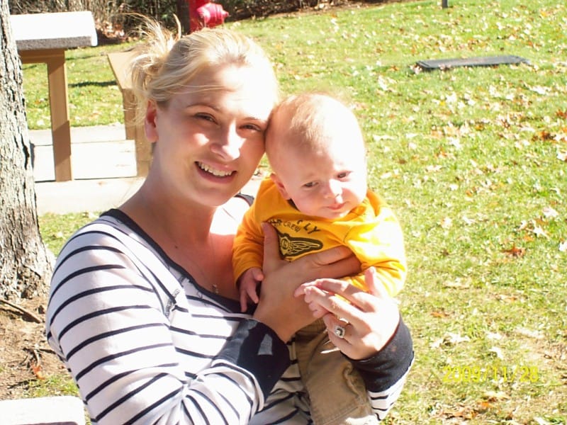 A woman with blonde hair wearing a striped shirt holds a baby in a yellow outfit while sitting outdoors on a sunny day, ensuring the child's protection under the warm sun.