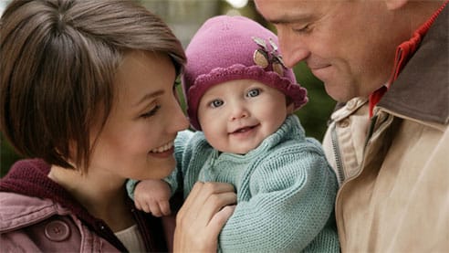 A woman and man smile at a baby wearing a purple hat, who is held between them and also smiling, enjoying the peace of mind brought by their no contract security system.