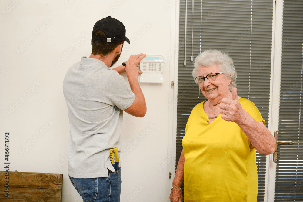 A technician in a gray shirt installs a security keypad on a wall while an elderly woman wearing a yellow shirt stands nearby, giving a thumbs-up gesture. It's clear that Atlanta's home security systems are bringing peace of mind to the community.