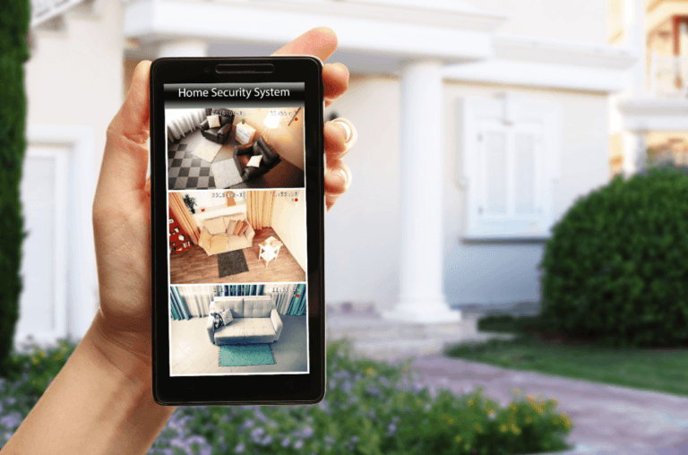 A hand holds a smartphone displaying a home security system app with live camera feeds from two indoor locations. A house and garden are visible in the background, showcasing the advanced features of East Cobb's premier home security systems.