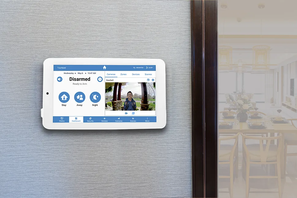 Switch Alula Security Systems: A smart security panel from Alula security systems mounted on a wall displays a touchscreen interface with options for stay, away, and night modes along with a live video feed. A dining area is visible in the background.