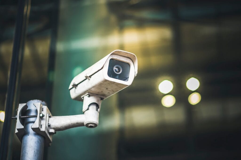 A close-up view of a white, outdoor security camera mounted on a pole, with a blurred reflective background.