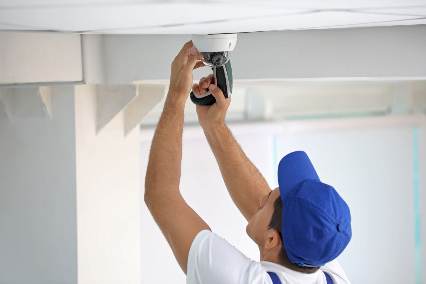 A person in a blue cap and white shirt completes the installation of a CCTV security camera on the ceiling.