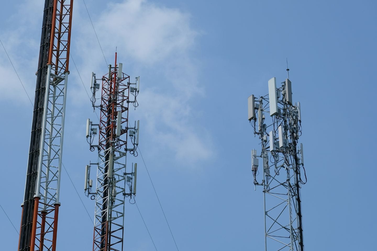 Three communication towers of varying heights stand against a blue sky with some clouds, featuring multiple antennas and equipment.