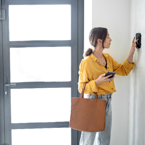 A woman in a yellow shirt and jeans activates an existing system using a wall-mounted device near the door, holding her phone and bag.