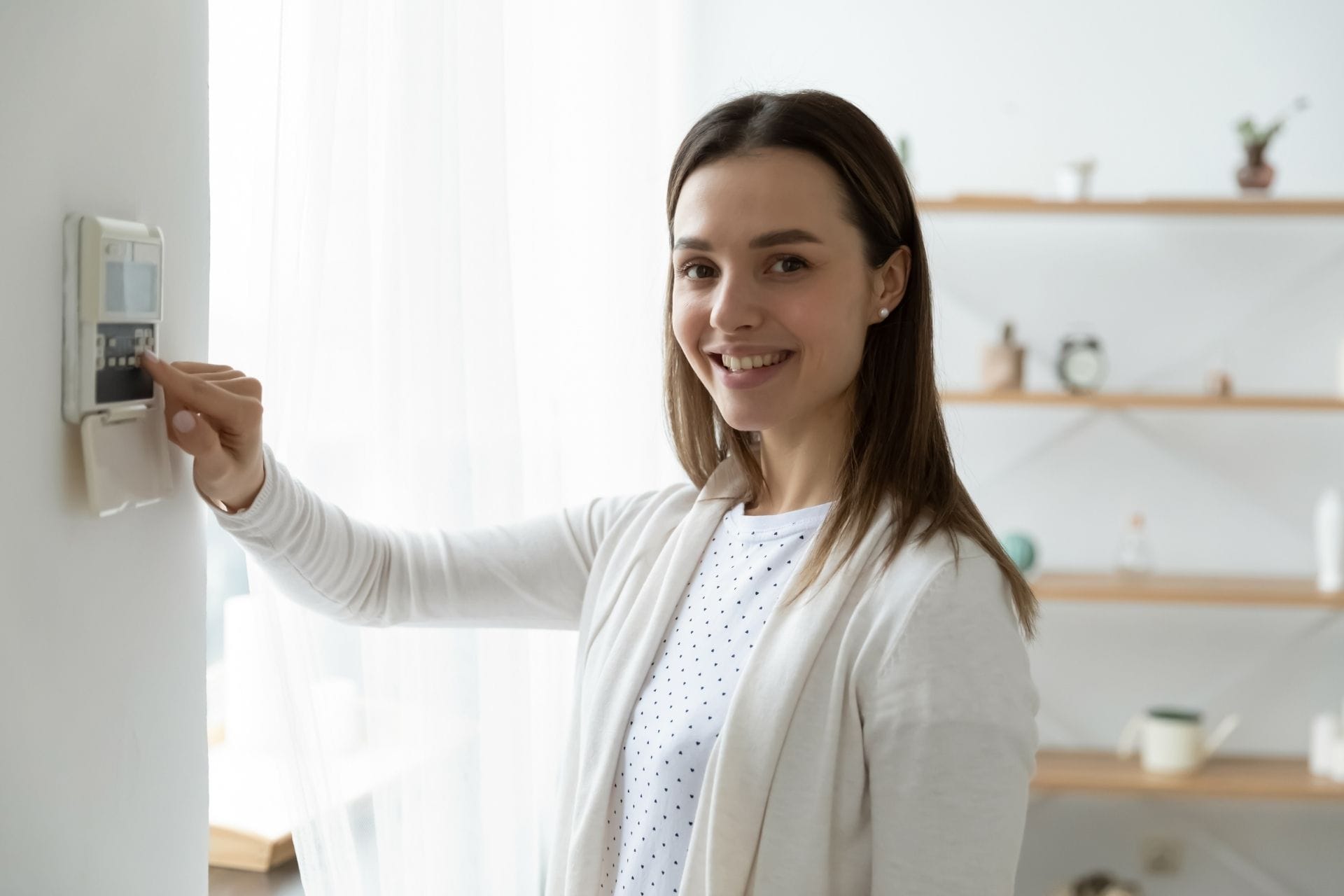 A woman adjusts a wall-mounted thermostat with a smile in a brightly lit room. Shelves with decorative items and discreet home security devices are in the background.