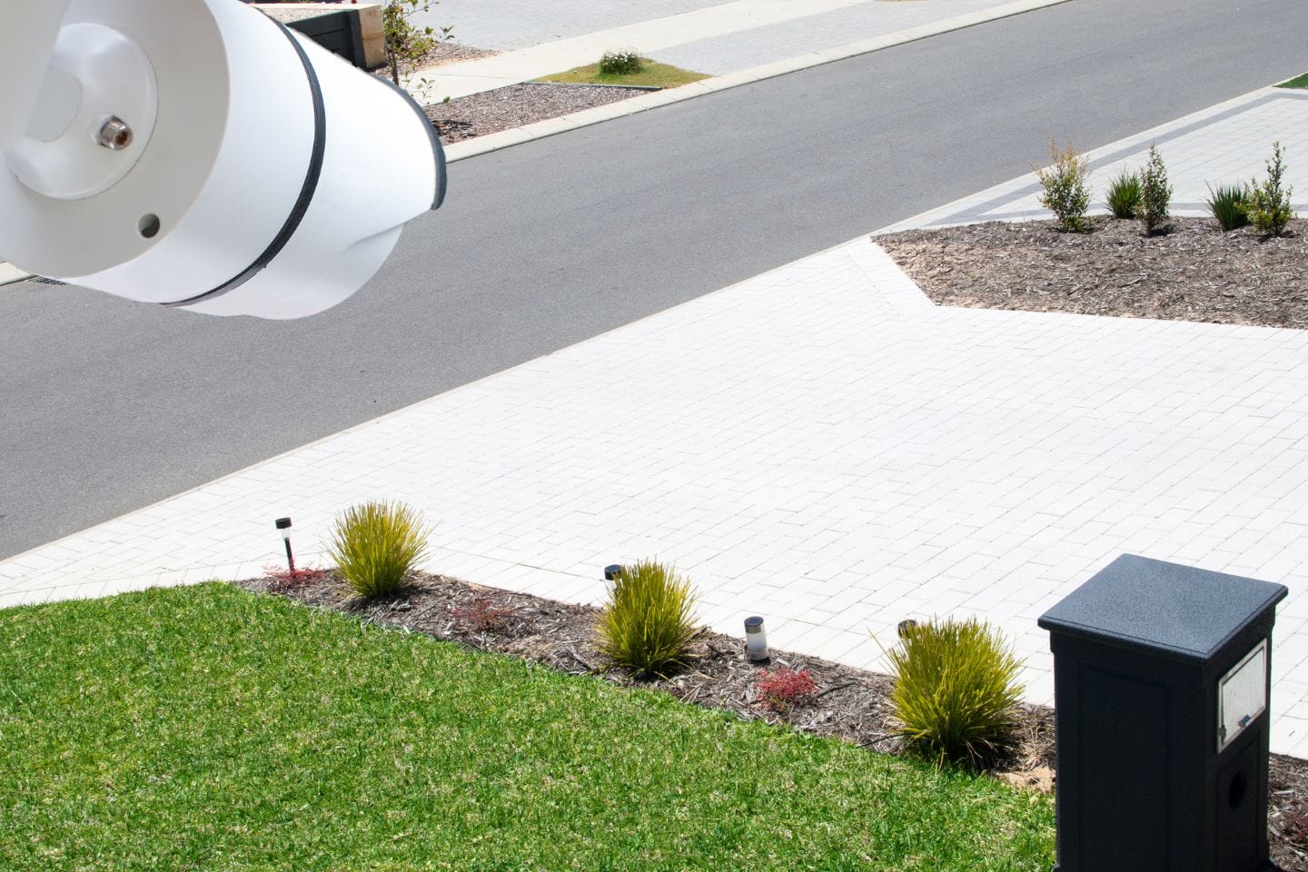 A security camera, part of the Total Connect system from Resideo, is positioned above a suburban street. The foreground shows a neatly manicured lawn and a black mailbox, while the background reveals a paved driveway and road.