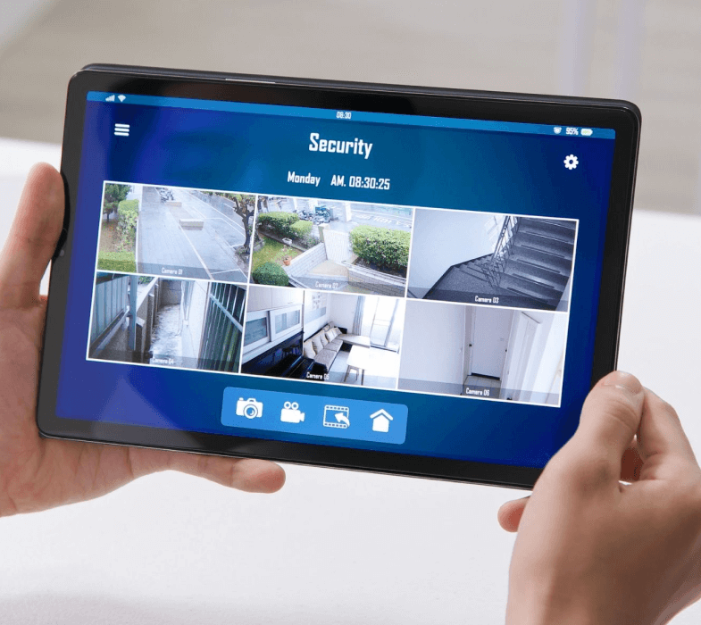 Person holding a tablet displaying a home security app interface with various camera feeds, date and time information, and various control icons on the screen.