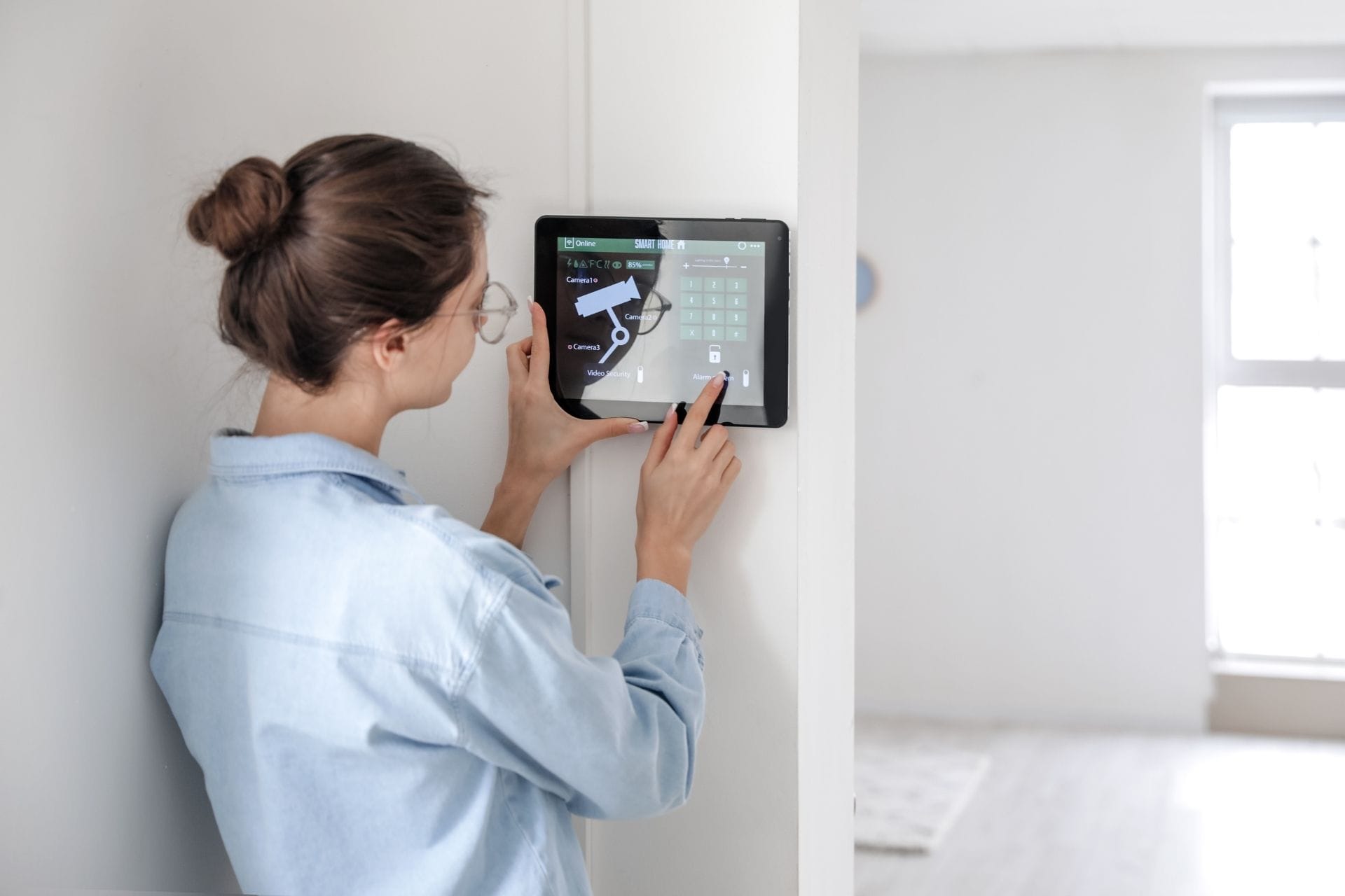 A person in a denim shirt uses a wall-mounted tablet to control a home security system, ensuring home safety inside a bright, modern room.