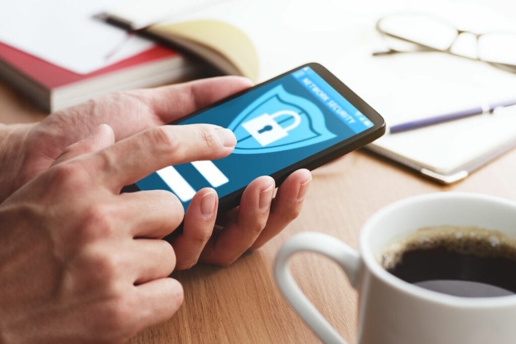 A person uses a smartphone with a security lock screen displayed at home. A cup of coffee, notebook, and pen are on the table.