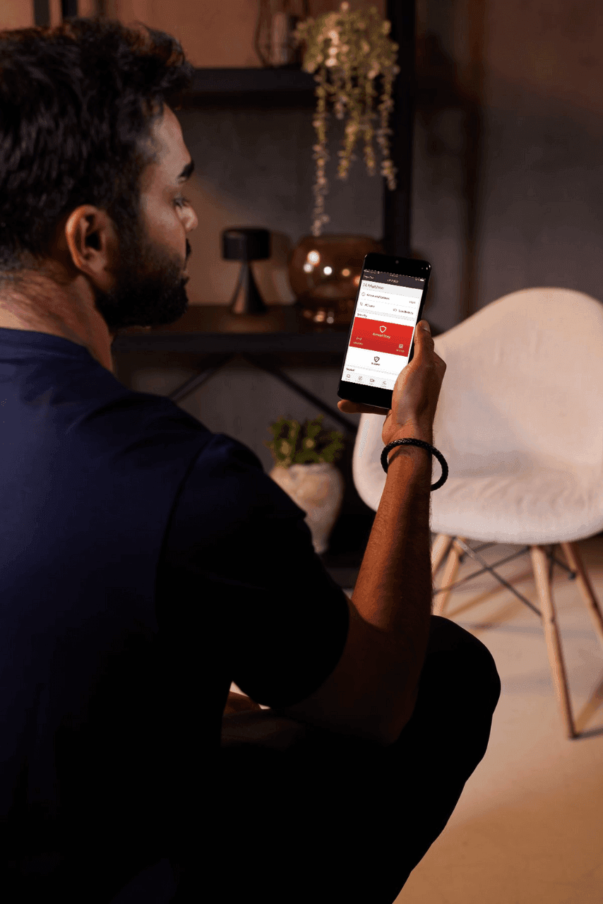 A man sits in a room, holding and looking at a smartphone with the Honeywell Total Connect app open on the screen. A white chair and a shelf with various items are in the background.