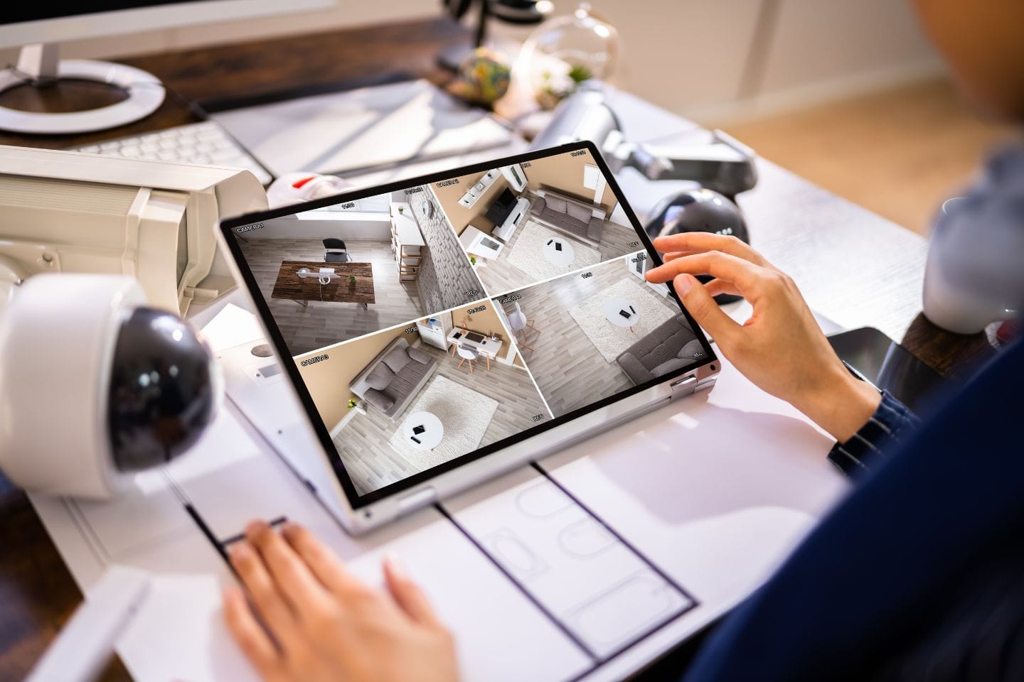 Person views surveillance footage of a home interior on a tablet, with four camera feeds shown. Various security devices and business office supplies are visible on the desk.