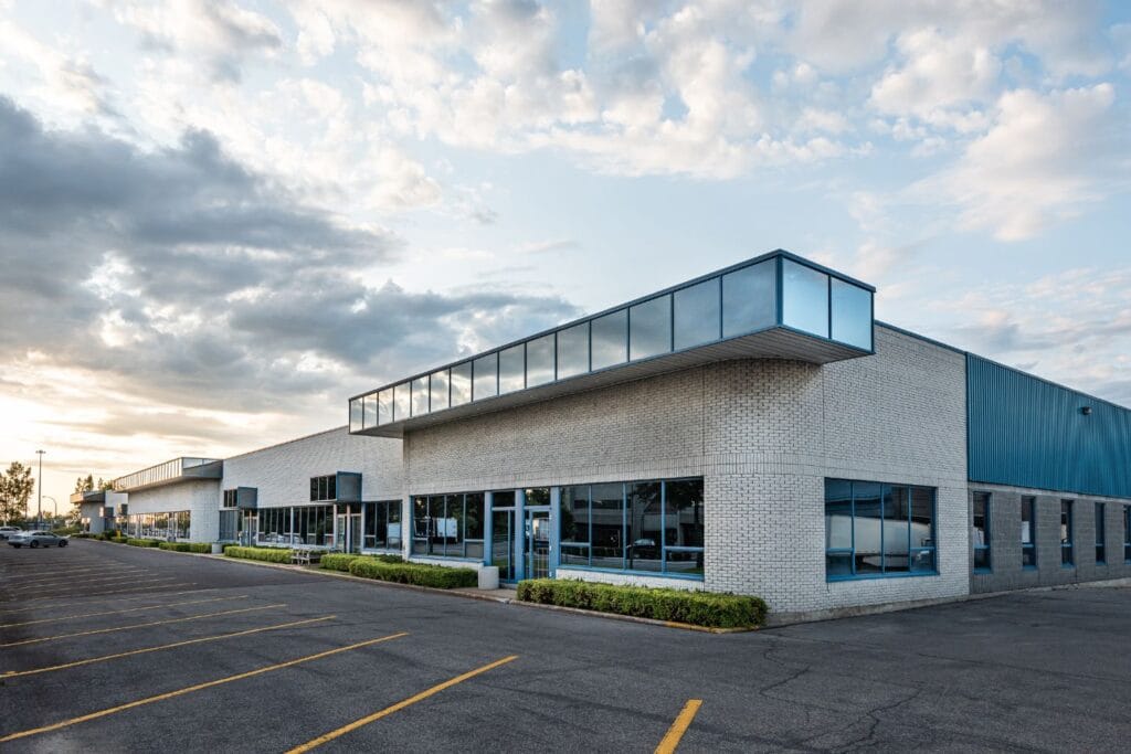 A modern industrial building with a white facade and large glass windows, featuring advanced business security systems, is located next to an empty parking lot under a partly cloudy sky.