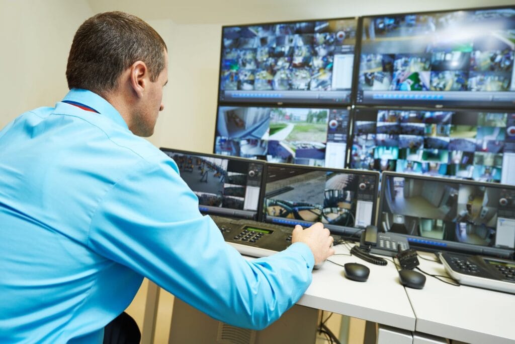 A man in a blue shirt monitors multiple security camera feeds on several screens at a security control station, ensuring the business operations remain safe and secure.