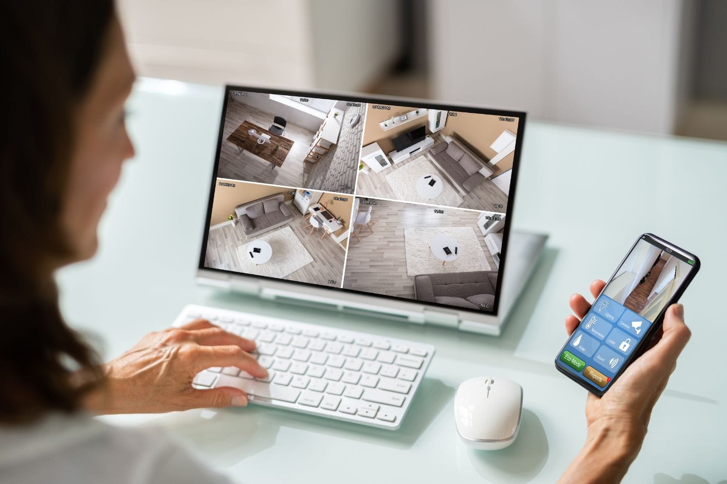 A person monitoring a home security system on a desktop computer screen with four camera views, while holding a smartphone showing a business security app interface.