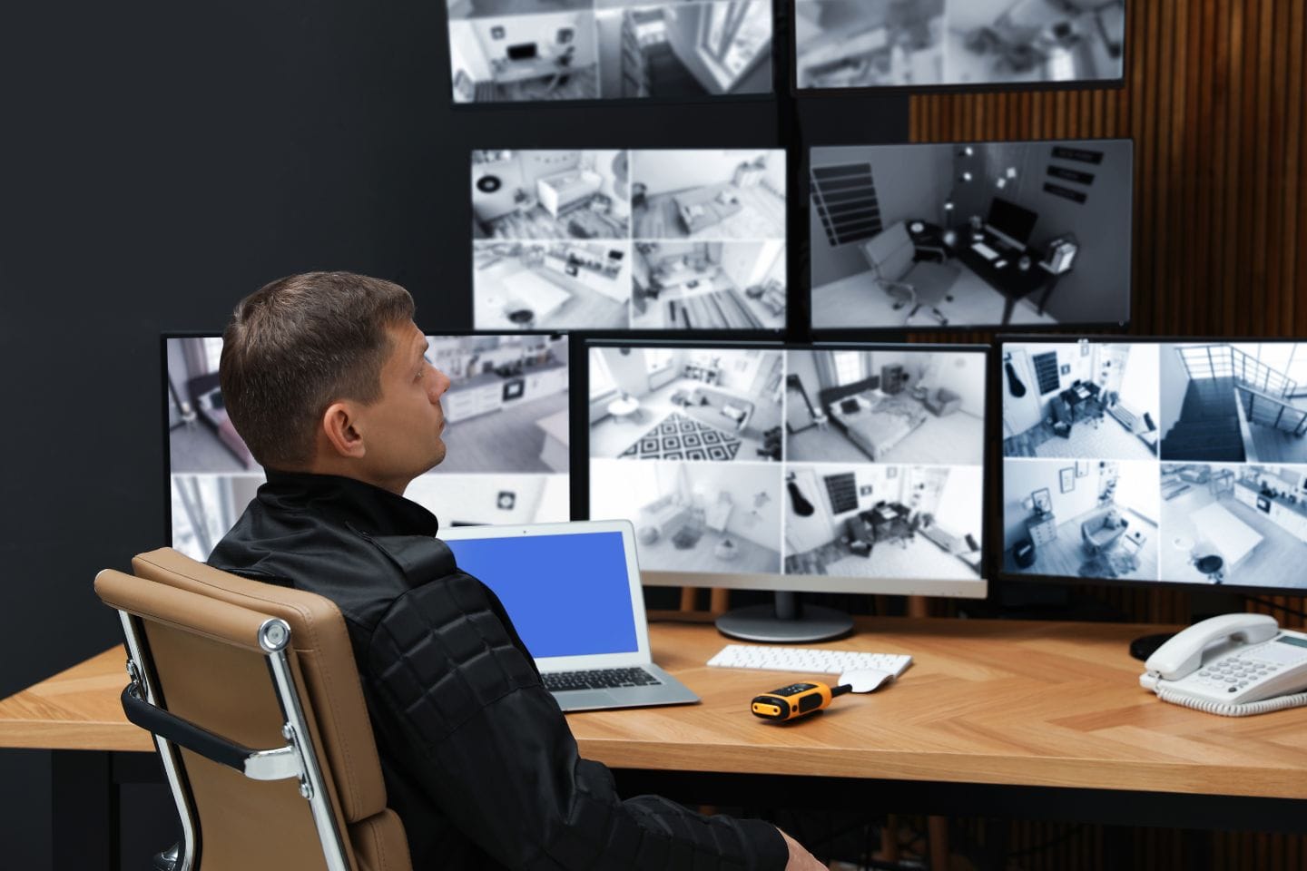 A person sits at a desk with multiple surveillance monitors, a laptop, and a digital handheld device, diligently observing security camera footage to ensure business security.