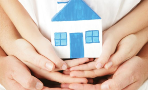 Multiple hands are cupped together holding a small, white house model with a blue roof and windows.