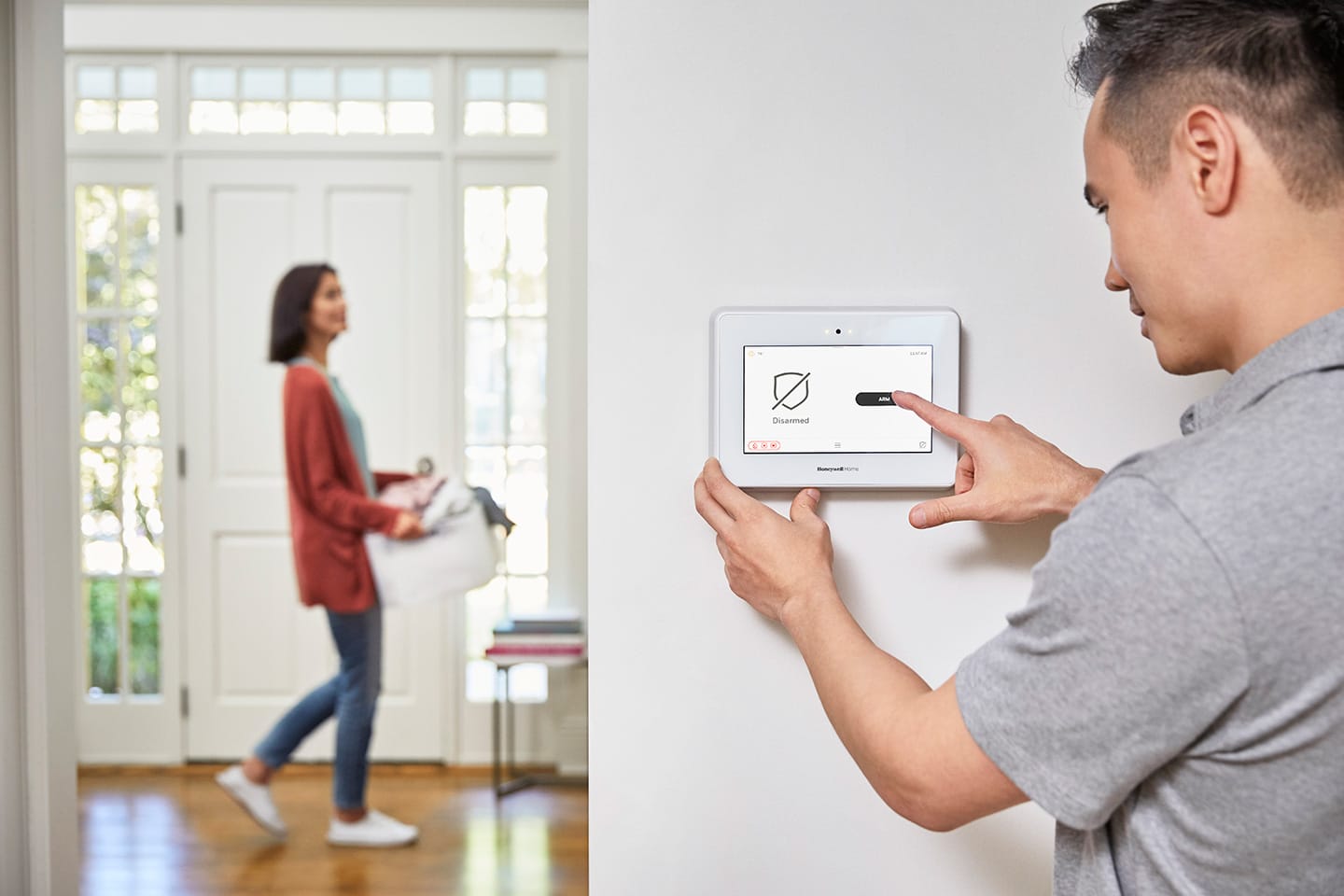 A person adjusts a smart home control panel on the wall, ensuring optimal home security, while another person walks by holding towels.